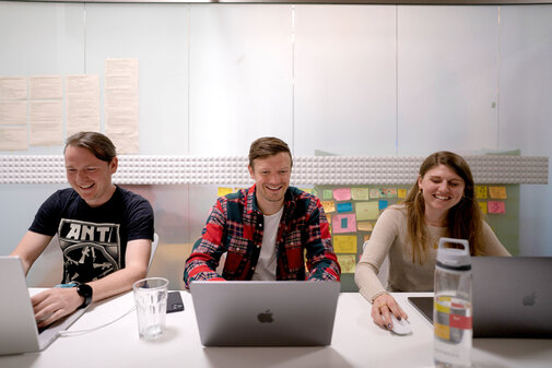 Three people laughing while working on IT projects on their laptops
